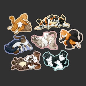 May include: A collection of seven die-cut stickers featuring pairs of cats in various playful wrestling poses. The cats are depicted in diverse coat patterns including orange tabby, calico, black, white, and brown tabby, all rendered in a cartoon style.