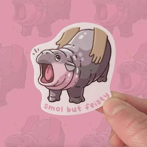 May include: A die-cut sticker showing a cartoon hippo with an open mouth. The hippo is grey with pink details and the text reads "Smol but feisty". The sticker is held by a hand against a pink background.
