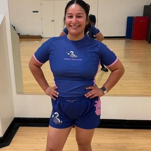 May include: A blue athletic top and shorts set with pink accents. The top and shorts feature the text "SWEATY ROLLS CLUB" and a cartoon illustration of a swan. The outfit is designed for athletic activities.