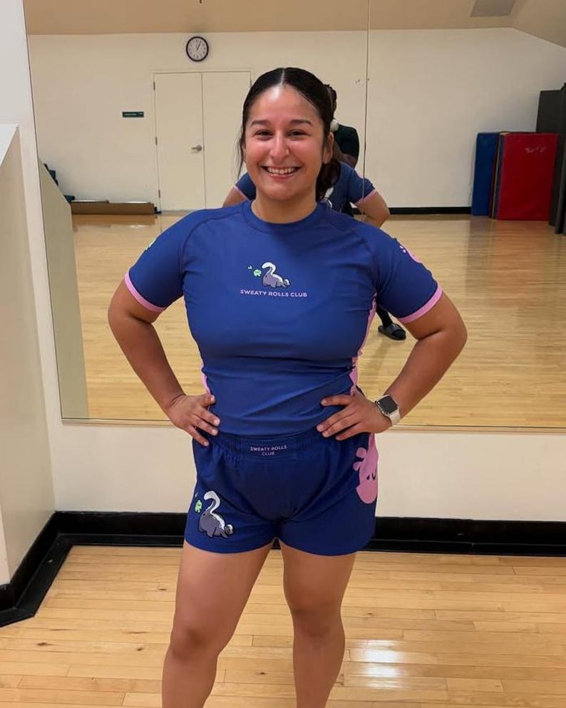 May include: A blue athletic top and shorts set with pink accents. The top and shorts feature the text "SWEATY ROLLS CLUB" and a cartoon illustration of a swan. The outfit is designed for athletic activities.