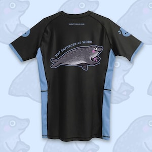 Op de afbeelding: Zwart sportshirt met lichtblauwe accenten en een cartoon zeehond ontwerp. De tekst "MAT ENFORCER AT WORK" staat onder de zeehond. De woorden "SWEATY ROLLS CLUB" staan op de achterkant van het shirt.