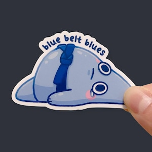 May include: A die-cut sticker featuring a sad, light blue, blob-like character wearing a dark blue martial arts belt. The character has large eyes, pink cheeks, and a tear. The text "blue belt blues" is printed in dark blue above the character.