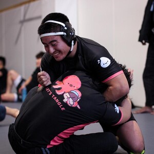 May include: Two people grappling in a gym, wearing black rash guards and headgear. One shirt has a pink octopus graphic and the text "LET'S TANGLE". The other shirt has a white logo on the sleeve. The background is blurred.