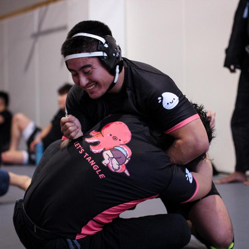 May include: Two people grappling in a gym, wearing black rash guards and headgear. One shirt has a pink octopus graphic and the text "LET'S TANGLE". The other shirt has a white logo on the sleeve. The background is blurred.