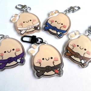 Puede incluir: Cinco llaveros de acrílico que presentan un personaje redondo, beige y sonriente con mejillas rosadas. Cada personaje lleva un cinturón de artes marciales de diferente color: blanco, azul, morado, marrón y negro con una punta roja.