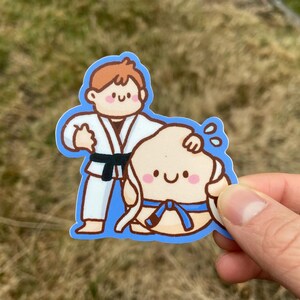 Peut inclure: Un autocollant représentant deux personnages de dessins animés en tenues d'arts martiaux. Un personnage, en kimono blanc avec une ceinture noire, aide un autre personnage, qui porte une ceinture bleue. L'autocollant a une bordure bleue et est tenu par une main.