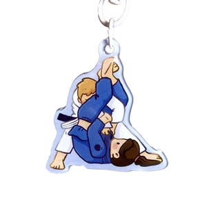 May include: A clear acrylic keyring featuring a cartoon illustration of two figures engaged in a grappling match. One figure wears a white gi with a black belt, while the other wears a blue gi. The figures are locked in a ground position.
