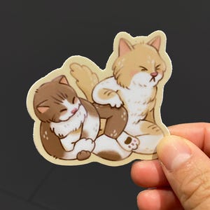 Op de afbeelding: Een gestanste sticker met een bruin-witte cyperse kat die opgerold slaapt naast een lichtoranje en witte kat die rechtop zit met gesloten ogen en een norse uitdrukking op het gezicht, weergegeven in een cartoon-kunststijl.