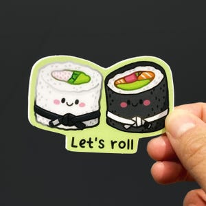 Peut inclure: Un autocollant découpé représentant deux sushis en dessin animé avec des visages souriants, l'un blanc avec une ceinture noire et l'autre noir avec une ceinture blanche, garnis d'avocat vert et de saumon orange, avec le texte Let's roll en noir.
