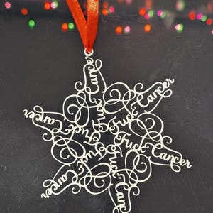 May include: A white, snowflake-shaped ornament with the words "Fuck Cancer" in a repeating pattern. The ornament is suspended by a red ribbon against a dark background with blurred, colorful lights.