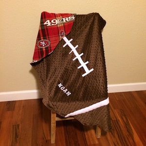 May include: A brown football-shaped blanket with white stitching and the name "NOAH" embroidered on it. The blanket has a red plaid section with the "49ERS" logo. The blanket is draped over a small wooden stool.