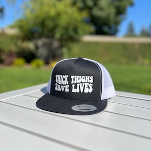 May include: Black and white trucker hat with the text "THICK THIGHS SAVE LIVES" in white retro font.