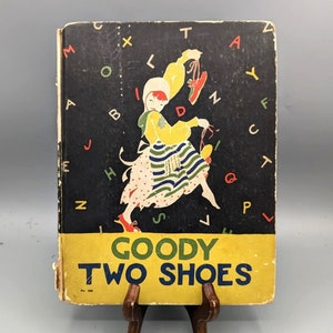 Könnte beinhalten: Ein Vintage-Kinderbuchcover mit gelbem Hintergrund und dem Titel "Goody Two Shoes" in grünen Buchstaben. Das Cover zeigt ein Mädchen in einem gestreiften Kleid und roten Schuhen, das auf einem schwarzen Hintergrund mit bunten Buchstaben steht.