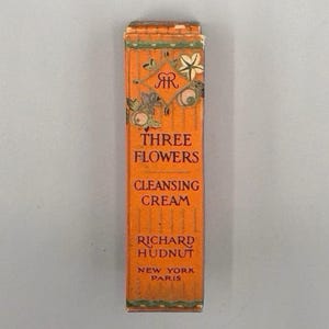 Op de afbeelding: Een oranje doos van "THREE FLOWERS CLEANSING CREAM" van Richard Hudnut, met bloemen en decoratieve accenten. De doos heeft een rechthoekige vorm en bevat tekst in vintage stijl, met de woorden "NEW YORK PARIS" onderaan.