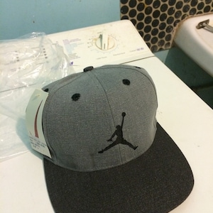 Puede incluir: Una gorra de b&eacute;isbol gris y negra con un logotipo bordado en negro de un jugador de baloncesto. El logotipo es una silueta de una persona saltando con un bal&oacute;n de baloncesto.
