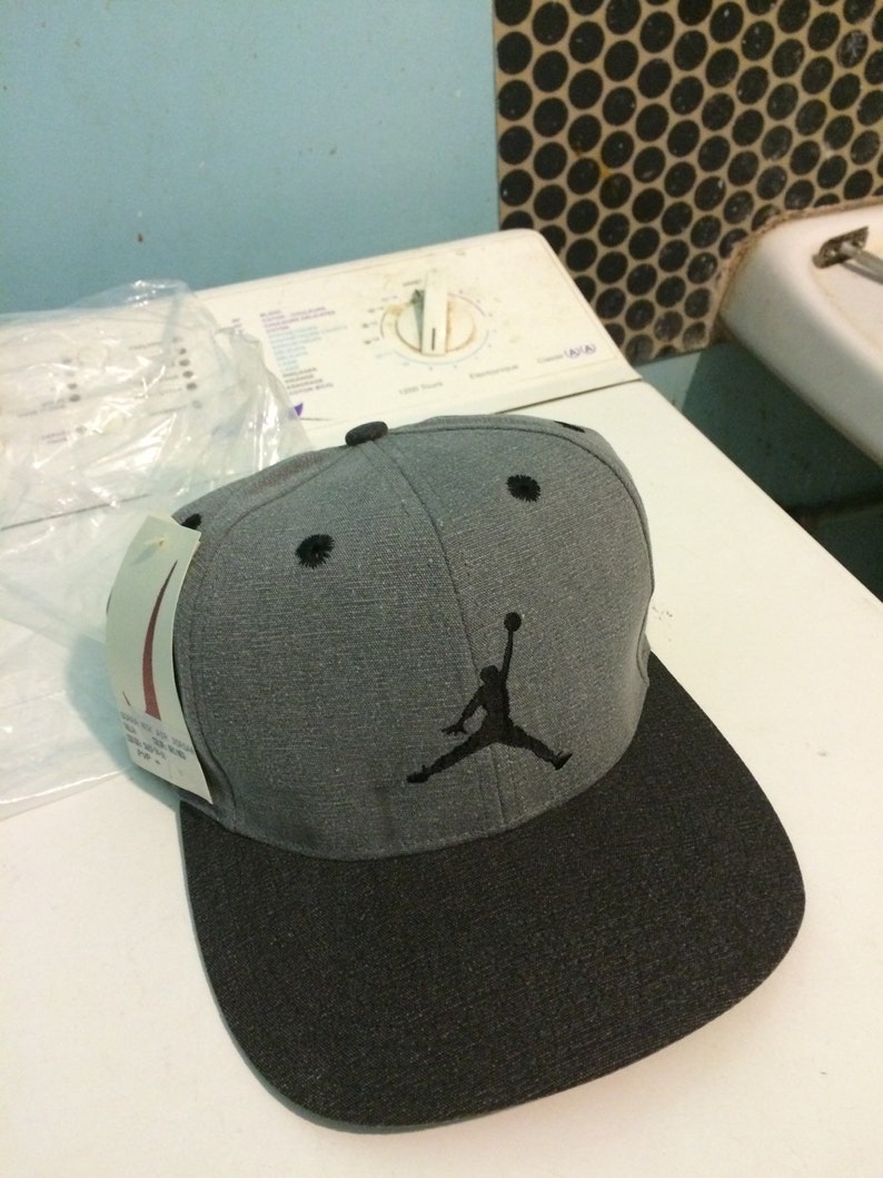Puede incluir: Una gorra de b&eacute;isbol gris y negra con un logotipo bordado en negro de un jugador de baloncesto. El logotipo es una silueta de una persona saltando con un bal&oacute;n de baloncesto.