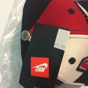 May include: A black, red and white baseball cap with a Nike logo on the front and a Nike tag attached. The tag has the text "NIKE" in white on a red background.