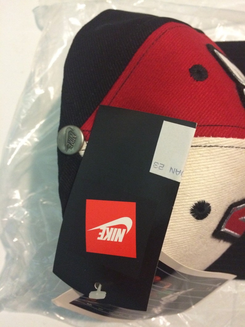 May include: A black, red and white baseball cap with a Nike logo on the front and a Nike tag attached. The tag has the text "NIKE" in white on a red background.