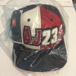 May include: A black, white and red baseball cap with the text "AJ23" in white letters on the front. The cap is in a plastic bag.