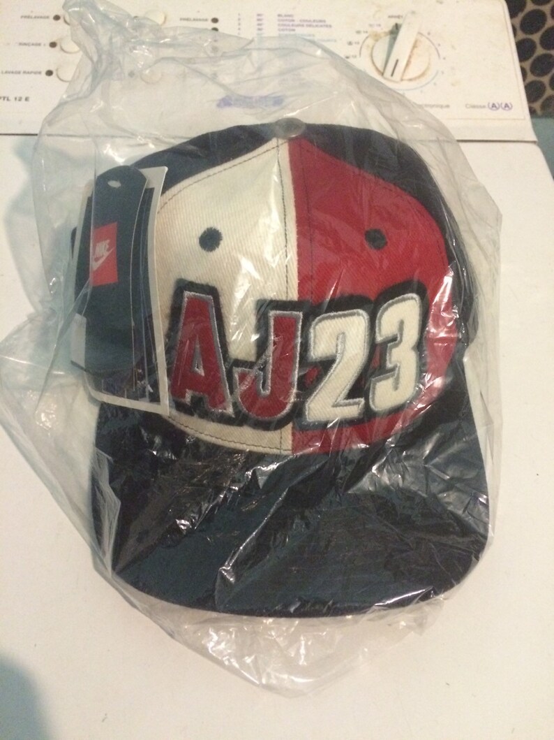 May include: A black, white and red baseball cap with the text "AJ23" in white letters on the front. The cap is in a plastic bag.