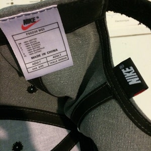 Puede incluir: Gorra de b&eacute;isbol gris con un logotipo de Nike negro y una correa ajustable negra. La etiqueta de la gorra dice "Nike" y "F50206 WDI" e indica que la gorra est&aacute; hecha en China.