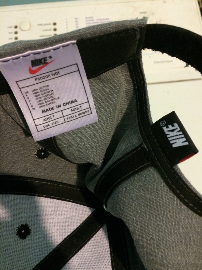 Puede incluir: Gorra de b&eacute;isbol gris con un logotipo de Nike negro y una correa ajustable negra. La etiqueta de la gorra dice "Nike" y "F50206 WDI" e indica que la gorra est&aacute; hecha en China.