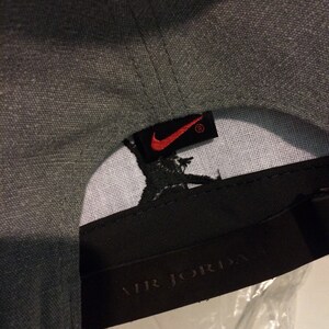 Puede incluir: Gorra de b&eacute;isbol gris con un logotipo de Nike negro y rojo y una etiqueta negra que dice "AIR JORDAN" en letras blancas.