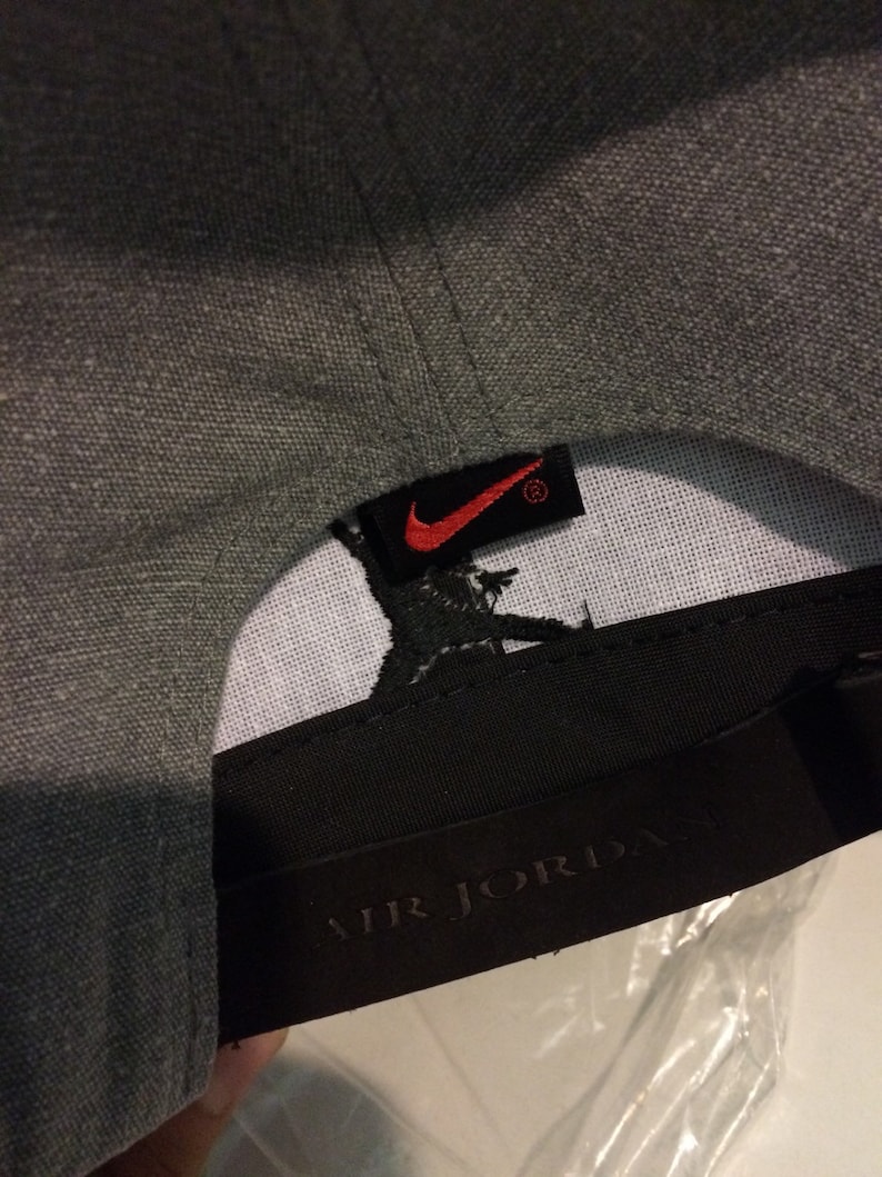 Puede incluir: Gorra de b&eacute;isbol gris con un logotipo de Nike negro y rojo y una etiqueta negra que dice "AIR JORDAN" en letras blancas.
