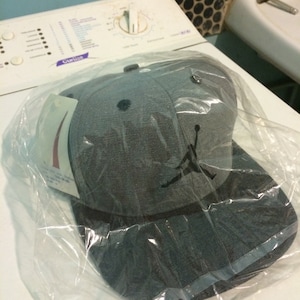 Puede incluir: Una gorra de b&eacute;isbol gris y negra con una silueta negra de un jugador de baloncesto en la parte delantera. La gorra est&aacute; en una bolsa de pl&aacute;stico.
