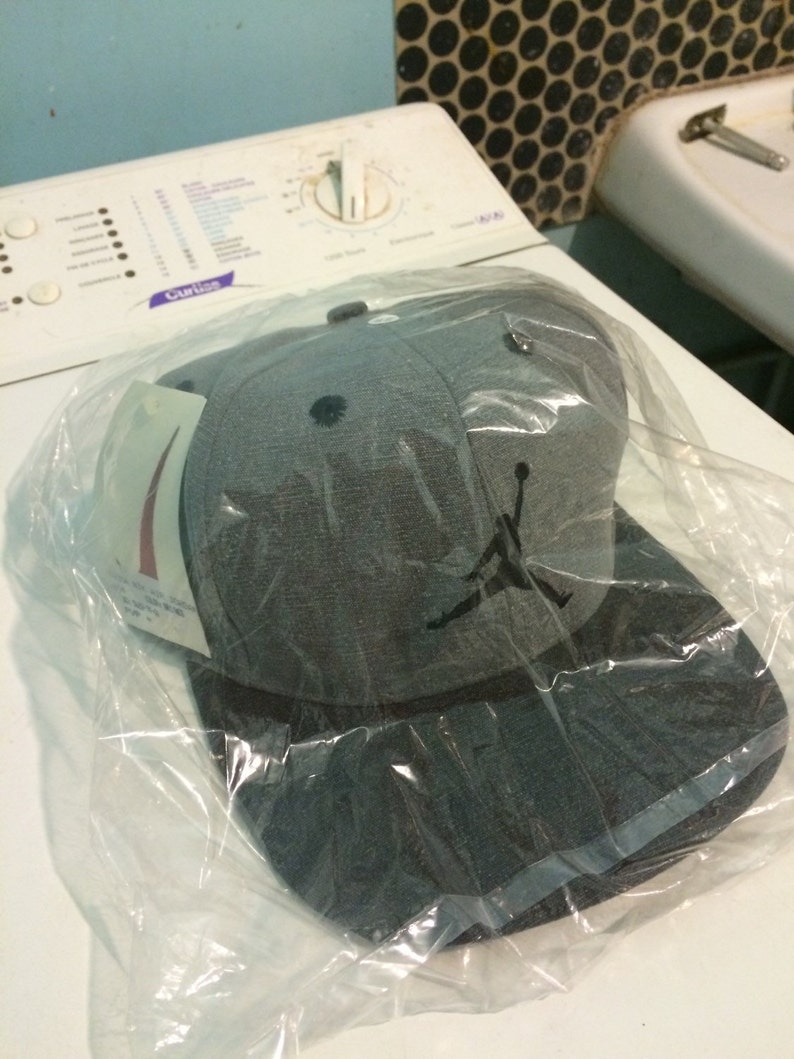 Puede incluir: Una gorra de b&eacute;isbol gris y negra con una silueta negra de un jugador de baloncesto en la parte delantera. La gorra est&aacute; en una bolsa de pl&aacute;stico.