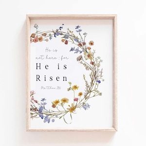 Pode incluir: Uma impressão emoldurada branca com uma grinalda floral feita de flores secas em tons de azul, amarelo e rosa. O texto "He is not here: for He is Risen. Matthew 28:6" está impresso em preto.