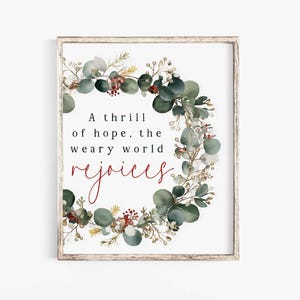 Puede incluir: Impresión enmarcada con una corona botánica de hojas de eucalipto verdes, bayas y ramitas. El texto "A thrill of hope, the weary world rejoices" se muestra en negro y rojo. El marco es de color claro.