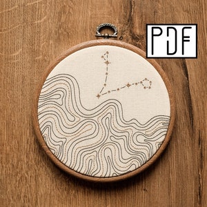 May include: A white embroidery hoop with a brown wooden frame. The embroidery features a brown and gold outline of the Pisces constellation with a white background.