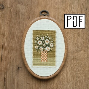 May include: Oval wooden embroidery hoop with a cross-stitch design of white flowers in a vase. The vase is orange and the background is olive green. The hoop has a metal clasp and the text "PDF" is in the upper right corner.
