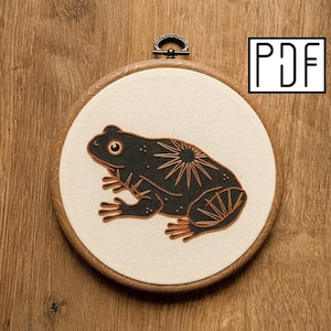 May include: An embroidery hoop with a black frog design and sunburst details in a wooden frame. The frog is on cream fabric. The text "PDF" is in the upper right corner. The hoop is hanging on a wooden surface.
