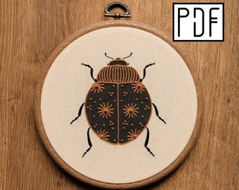 May include: A circular wooden embroidery hoop containing a cream-colored fabric with a hand-stitched black beetle design. The beetle features orange thread detailing in starburst and dot patterns. The letters PDF are printed in a black square box.