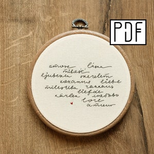 May include: Embroidered hoop with the words "amore", "láska", "mikość", "ljubezen", "szerelem", "кохання", "liebe", "milestība", "ränka", "kärlek", "liefde", "love", "amreur" in different languages. A small red heart is embroidered in the center.