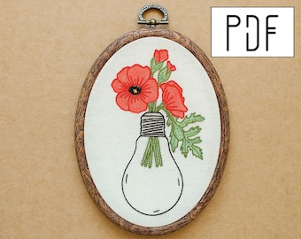 May include: An oval embroidery hoop containing white fabric with a stitched design of red poppies emerging from a light bulb. The hoop has a dark brown wood-grain frame and a metal hanging loop at the top. The text PDF is visible in a white box.