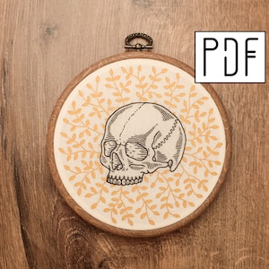 May include: A round embroidery hoop featuring a detailed black and white skull design. The skull is centered on a cream-colored fabric with a yellow leaf pattern. The hoop is made of wood and has a metal hanging loop. The letters "PDF" are in the upper right corner.
