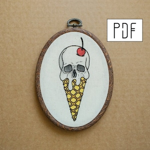 May include: Embroidered artwork in an oval wooden hoop. The design depicts a skull-shaped ice cream cone, with a cherry on top. The cone is yellow, and the skull is white with black eye sockets. The text "PDF" is visible.