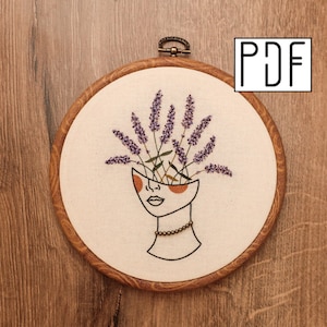 May include: Embroidered hoop art featuring a stylised face with lavender flowers growing out of the top of the head. The face is outlined in black thread and has orange circles for eyes. The lavender flowers are purple and green.
