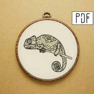 May include: Embroidered hoop art featuring a gray chameleon with floral patterns on its body, perched on a branch. The hoop is made of wood and has a brown finish.