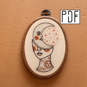 May include: An oval-shaped embroidery hoop featuring a celestial design. The artwork depicts a face split in two, with a crescent moon and stars above. The design includes a sun and cloud tattoo. The hoop has a dark wooden frame and a metal hanger.
