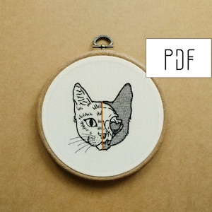 Pode incluir: Desenho bordado de uma cabeça de gato com um lado mostrando uma caveira e o outro lado mostrando um rosto de gato normal. O desenho está em um tecido branco esticado sobre um aro de madeira.