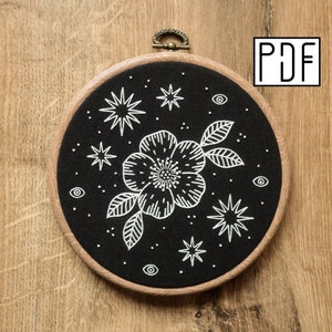 May include: Black embroidery hoop with a white floral design and stars. The design includes a large flower with leaves and small stars and eyes.