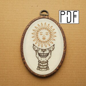 May include: Oval embroidery hoop featuring a sun design above a skull. The sun is golden with a serene face, and the skull is detailed in brown. The hoop has a wooden frame and a metal clasp at the top. The letters "PDF" are in the upper right corner.