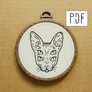 May include: A black and white embroidery pattern of a Sphynx cat with five eyes in a wooden embroidery hoop. The pattern is available as a PDF download.