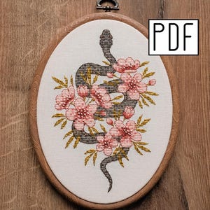 Peut inclure: Broderie au point de croix ovale représentant un serpent gris entouré de fleurs roses et de feuilles vertes. Le motif est encadré dans un cercle en bois avec un crochet en métal. Le texte "PDF" est en haut à droite.