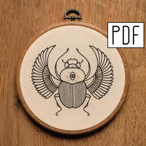 May include: Embroidered hoop art featuring a black line drawing of a scarab beetle with wings. The beetle has a large eye with a circle of small dots around it.