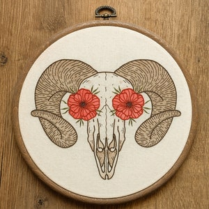 May include: A round, embroidered artwork featuring a ram skull with large, curved horns. The skull is cream-colored, with red flowers replacing the eye sockets. The artwork is set in a wooden hoop.